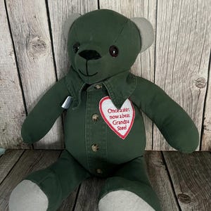 Op de afbeelding: Een knuffelbeer gemaakt van donkergroene stof met lichtgrijze oren en voetzolen. De beer heeft een knoopsluiting aan de voorzijde met bruine knopen en een witte hartvormige patch geborduurd met de rode tekst: Once a shirt now a bear, Grandpa Steel.