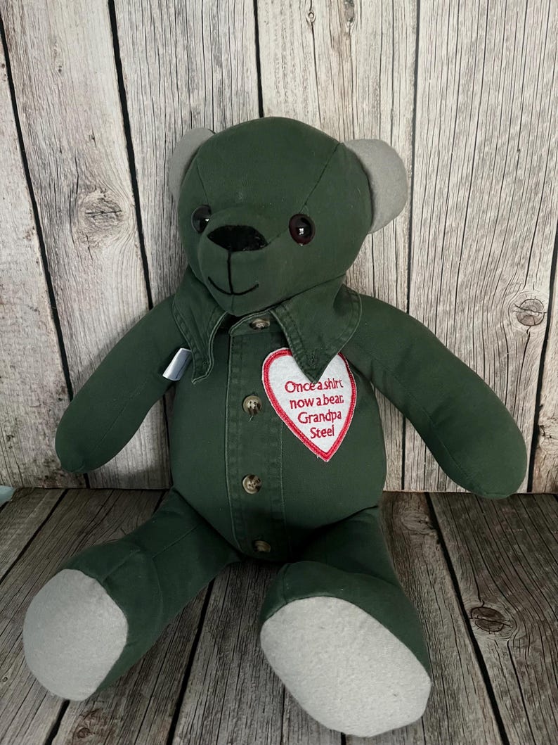 Op de afbeelding: Een knuffelbeer gemaakt van donkergroene stof met lichtgrijze oren en voetzolen. De beer heeft een knoopsluiting aan de voorzijde met bruine knopen en een witte hartvormige patch geborduurd met de rode tekst: Once a shirt now a bear, Grandpa Steel.