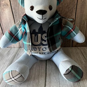 Op de afbeelding: Een knuffelbeer van lichtblauwe stof met donkerbruine ogen en een zwarte neus. De beer draagt een geruit overhemd in tinten van groenblauw, zwart en rood. Op de borst staat de tekst US MIDTOWN PIEDMONT PARK, ATLANTA. De oren en voeten hebben geruite accenten.