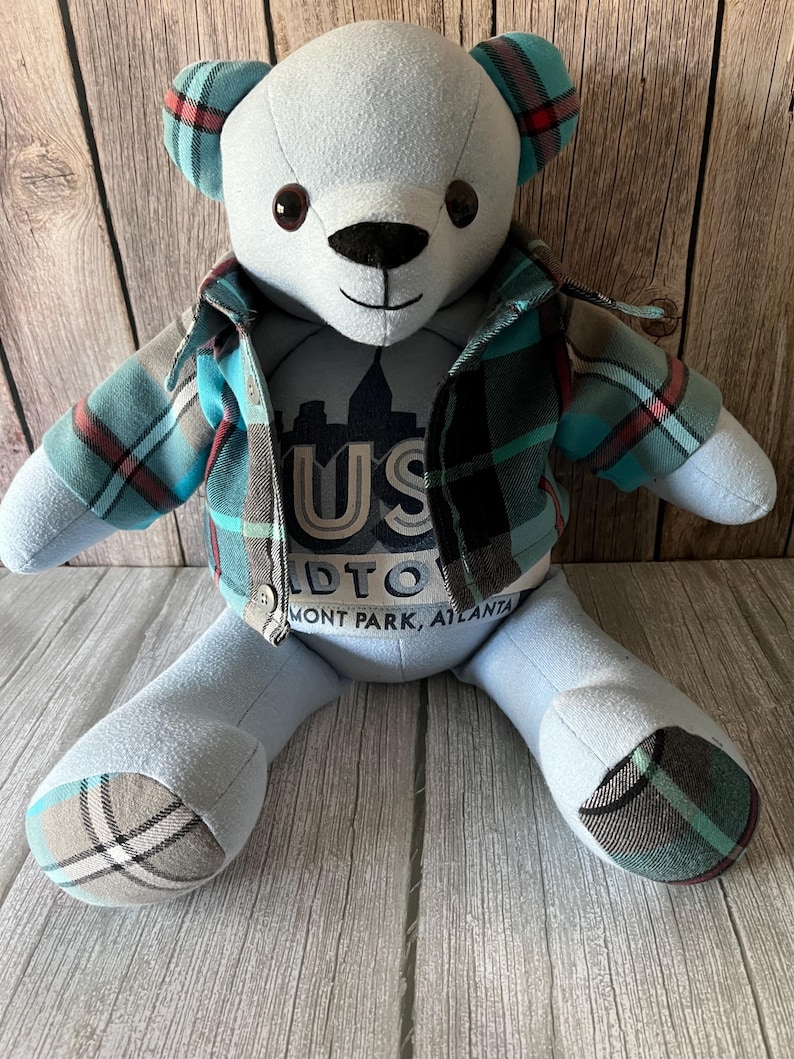 Op de afbeelding: Een knuffelbeer van lichtblauwe stof met donkerbruine ogen en een zwarte neus. De beer draagt een geruit overhemd in tinten van groenblauw, zwart en rood. Op de borst staat de tekst US MIDTOWN PIEDMONT PARK, ATLANTA. De oren en voeten hebben geruite accenten.
