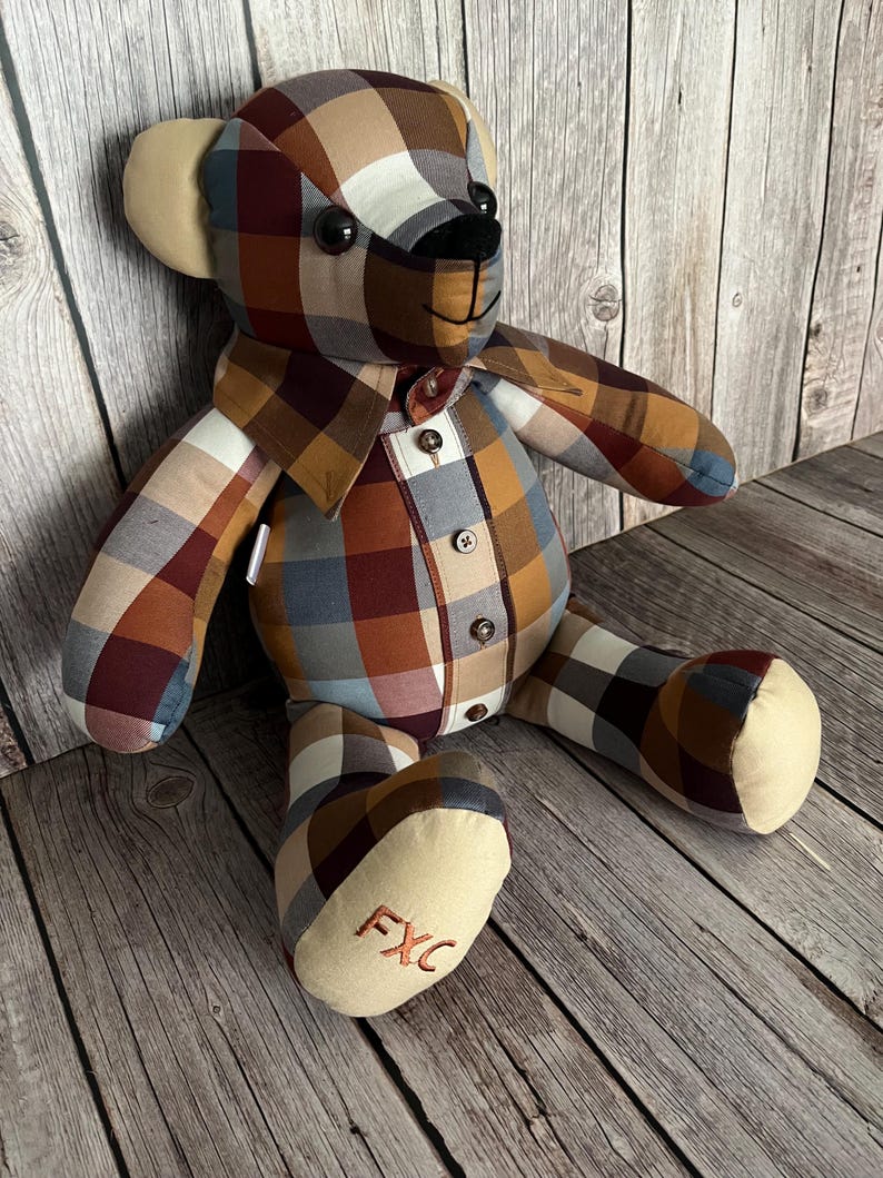 Op de afbeelding: Een knuffelbeer gemaakt van een geruit overhemd met een patroon van bruine, beige, blauwe en witte vierkanten. De beer heeft effen beige oren en voetzolen, zwarte knoopogen, een zwarte geborduurde neus en de geborduurde letters FXC op de voet.