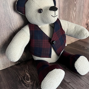 Op de afbeelding: Een knuffelbeer gemaakt van cr&egrave;mekleurige, gestructureerde stof. De beer draagt een vestje en beenstukken met een donkerrood en marineblauw ruitpatroon. Het heeft donkerbruine plastic ogen, een zwart gestikte neus en &eacute;&eacute;n zwarte knoop op het vest.