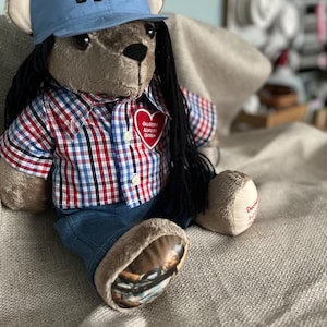 Op de afbeelding: Een pluche teddybeer met lang zwart haar, gekleed in een lichtblauwe honkbalpet met de tekst DBlock, een rood, wit en blauw geruit overhemd met een hartvormige patch en een blauwe spijkerbroek. Op &eacute;&eacute;n voet staat een afgedrukte foto.