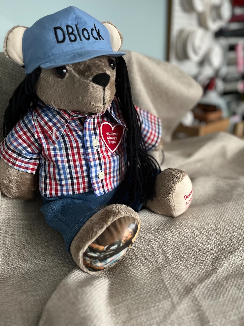Op de afbeelding: Een pluche teddybeer met lang zwart haar, gekleed in een lichtblauwe honkbalpet met de tekst DBlock, een rood, wit en blauw geruit overhemd met een hartvormige patch en een blauwe spijkerbroek. Op &eacute;&eacute;n voet staat een afgedrukte foto.