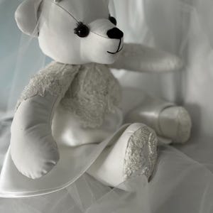 May include: A white teddy bear wearing a white satin dress with lace detailing and a white veil. The bear is sitting on a white fabric surface.