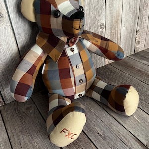 May include: A stuffed bear crafted from plaid fabric in shades of brown, tan, blue, and white. The bear features a button-down shirt front with a collar, dark round eyes, a black stitched nose, and the embroidered letters FXC on one foot.