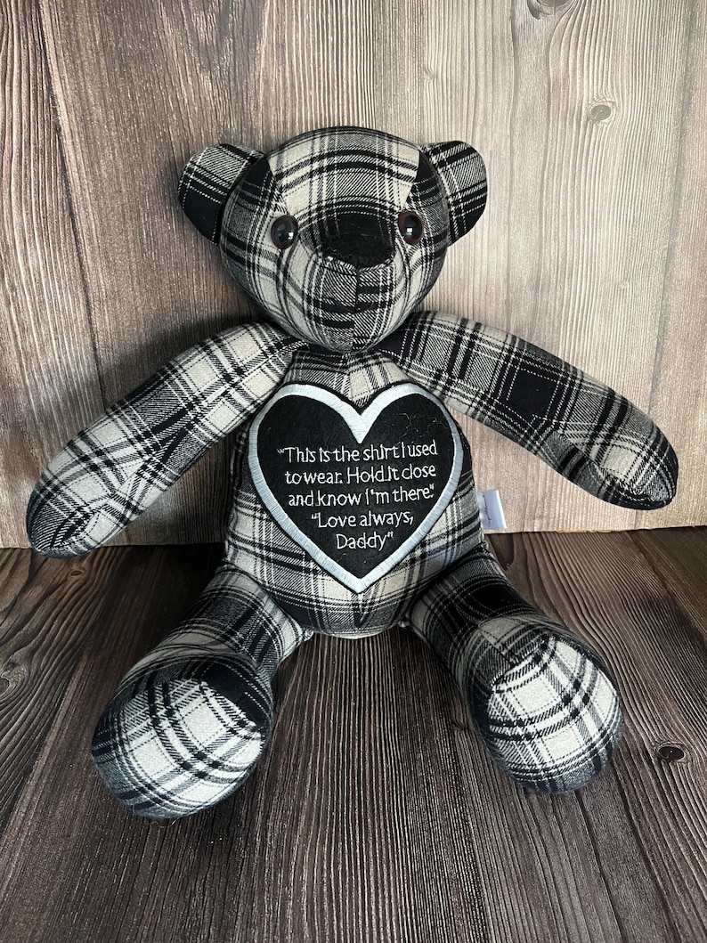 Op de afbeelding: Een knuffelbeer gemaakt van zwart, wit en grijs geruite stof. De beer heeft een zwart hartvormig embleem op de borst met witte geborduurde tekst: This is the shirt I used to wear. Hold it close and know I'm there. Love always, Daddy.