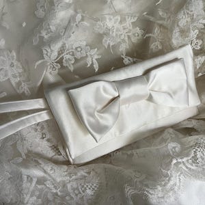 May include: A rectangular ivory satin clutch bag featuring a prominent centered bow on the front flap and a matching thin wrist strap. The clutch is crafted from a smooth, lustrous fabric and displays a clean, structured design with finished edges.