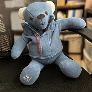 Op de afbeelding: Een blauwe pluche teddybeer met een blauw vest met rits, een geribbelde kraag en oranje accenten. De beer heeft lichtgekleurde oren en donkere ogen. Op de rechtervoet van de beer is de tekst "Dad 7-1-51" in wit garen geborduurd.