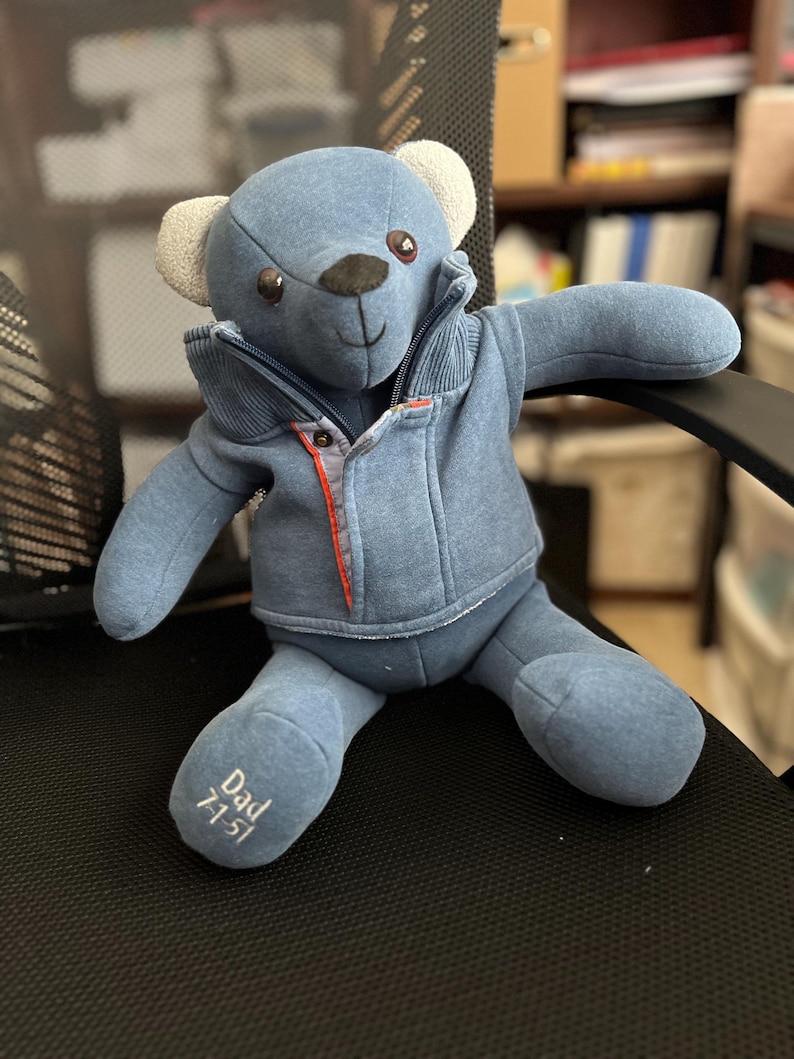 Op de afbeelding: Een blauwe pluche teddybeer met een blauw vest met rits, een geribbelde kraag en oranje accenten. De beer heeft lichtgekleurde oren en donkere ogen. Op de rechtervoet van de beer is de tekst "Dad 7-1-51" in wit garen geborduurd.