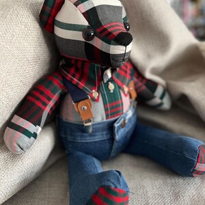 Op de afbeelding: Een knuffelbeer gemaakt van stof met een rood, groen, zwart en wit ruitpatroon. De beer draagt een geruit overhemd met een kraag en knopen, en een blauwe spijkerbroek die wordt opgehouden door bruine leren bretels met zilverkleurige gespen.