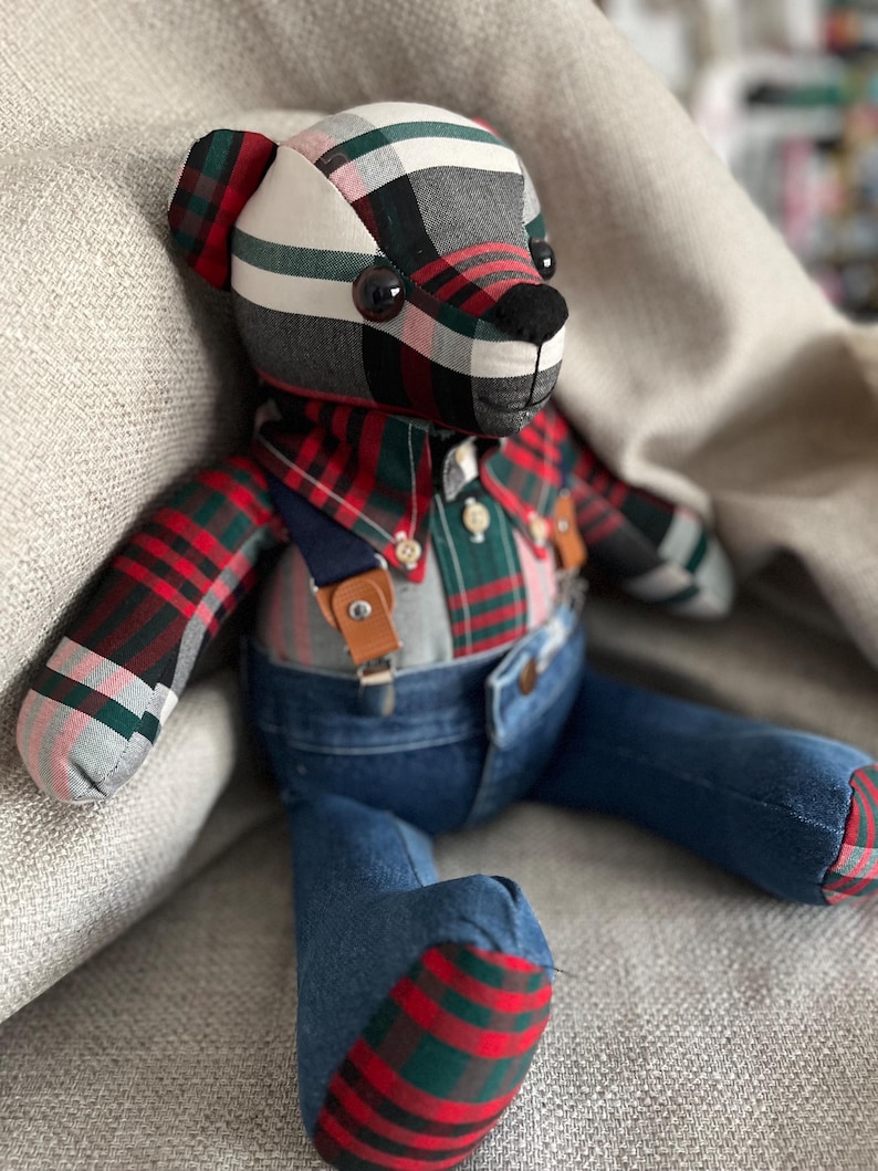 Op de afbeelding: Een knuffelbeer gemaakt van stof met een rood, groen, zwart en wit ruitpatroon. De beer draagt een geruit overhemd met een kraag en knopen, en een blauwe spijkerbroek die wordt opgehouden door bruine leren bretels met zilverkleurige gespen.