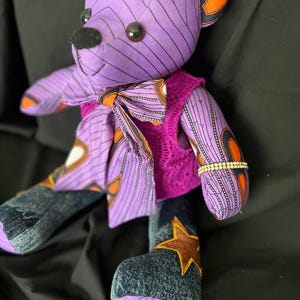 Op de afbeelding: Een knuffelbeer gemaakt van paarse stof met zwarte geometrische patronen en oranje accenten. Het draagt een bijpassende vlinderdas, een felroze kanten vestje en donkere denim benen met een bruine sterapplicatie. Een strass-armband siert een van de armen.