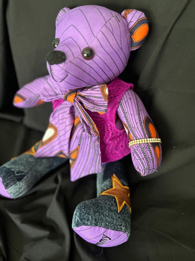 Op de afbeelding: Een knuffelbeer gemaakt van paarse stof met zwarte geometrische patronen en oranje accenten. Het draagt een bijpassende vlinderdas, een felroze kanten vestje en donkere denim benen met een bruine sterapplicatie. Een strass-armband siert een van de armen.