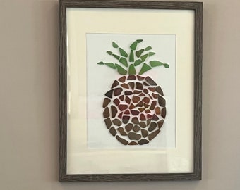Può includere: Un'opera d'arte incorniciata che raffigura un ananas realizzato con pezzi di vetro colorati. L'ananas è marrone con foglie verdi e si trova su uno sfondo bianco.