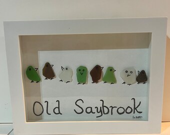 Può includere: Un'opera d'arte incorniciata che presenta una fila di otto piccoli uccelli di vetro in vari colori, tra cui verde, marrone, bianco e nero. Gli uccelli sono disposti su uno sfondo bianco con il testo "Old Saybrook" scritto sotto.