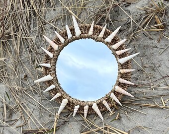 Può includere: Uno specchio rotondo con una cornice intrecciata realizzata con materiali naturali. La cornice è decorata con conchiglie bianche disposte a forma di sole.