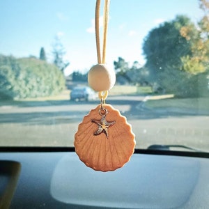 May include: A beige seashell car air freshener with a gold starfish charm hanging from a white string. The air freshener is hanging from the rearview mirror of a car.