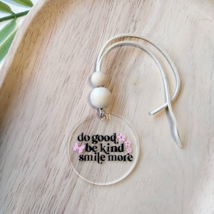 May include: A clear acrylic charm with the words "do good, be kind, smile more" in black script, decorated with pink flowers and a butterfly. It hangs from a white cord with two wooden beads.