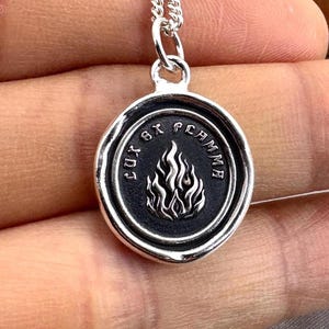 May include: A silver pendant necklace with a circular charm. The charm features a flame design in the center, surrounded by the words "Lux Ex Flamma". The necklace has a delicate chain.