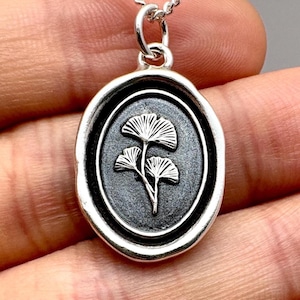 May include: Silver-toned pendant necklace featuring an oval-shaped pendant with a detailed ginkgo leaf design. The pendant has a textured, dark background and a polished silver border. The necklace chain is a delicate, twisted style.