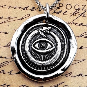 Può includere: Collana con pendente argentato raffigurante un ouroboros che circonda un occhio. Il pendente circolare ha un bordo strutturato ed è appeso a una catena. Lo sfondo è una carta vintage con testo manoscritto.