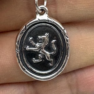 May include: Silver-toned pendant with an oval, dark background. A detailed, raised image of a heraldic lion is in the center. The pendant has a decorative border and a small loop for a chain.