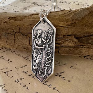 May include: A silver pendant necklace featuring a detailed relief of a classical figure holding a cup and a serpent. The pendant is rectangular with a pointed bottom and hangs from a delicate chain. The background includes aged paper and a wooden element.