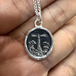 Può includere: Una collana d'argento con un ciondolo rotondo che presenta un'immagine dettagliata di un faro in un mare tempestoso. Il ciondolo è in stile sigillo di cera.