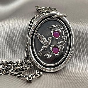 May include: A silver necklace with a pendant featuring a hummingbird in flight with two pink gemstones. The pendant is oval shaped and has a textured surface. The chain is a silver curb chain.
