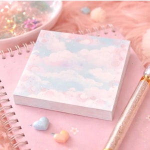 May include: A pad of square sticky notes, featuring a pastel sky and cloud design, sits on a pink spiral-bound notebook. A glittery pen and heart-shaped decorations are also present. The scene is set on a fluffy pink surface.