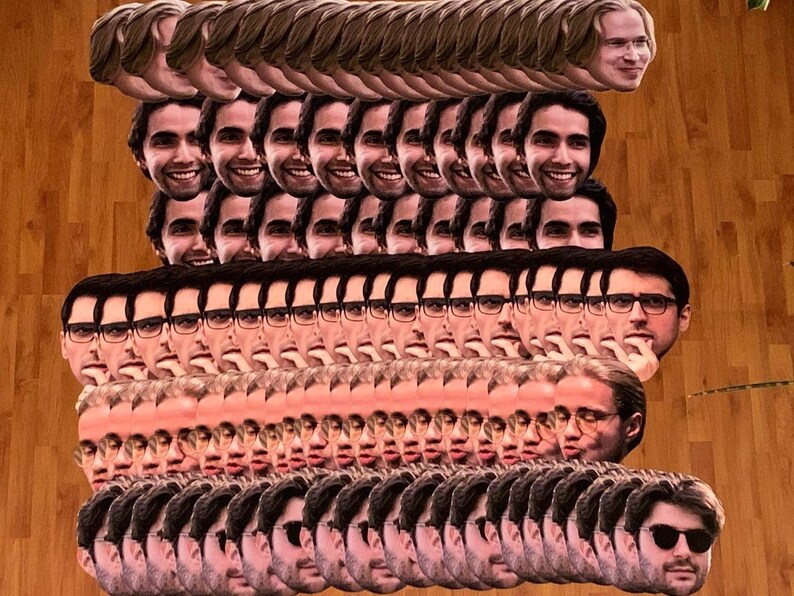 May include: A large stack of paper face masks featuring a man's face. The masks are arranged in rows and columns, with the man's face facing forward in each mask. The masks are printed on white paper and have a realistic appearance.
