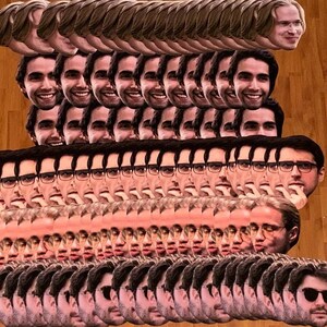 May include: A large stack of paper face masks featuring a variety of different people. The masks are all printed with realistic images of faces, some with glasses and some without.