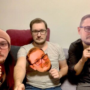 May include: Three people are holding up cardboard cutouts of their faces. The person on the left is wearing a brown beanie and a black shirt. The person in the middle is wearing a gray shirt and jeans. The person on the right is wearing a black shirt and jeans.