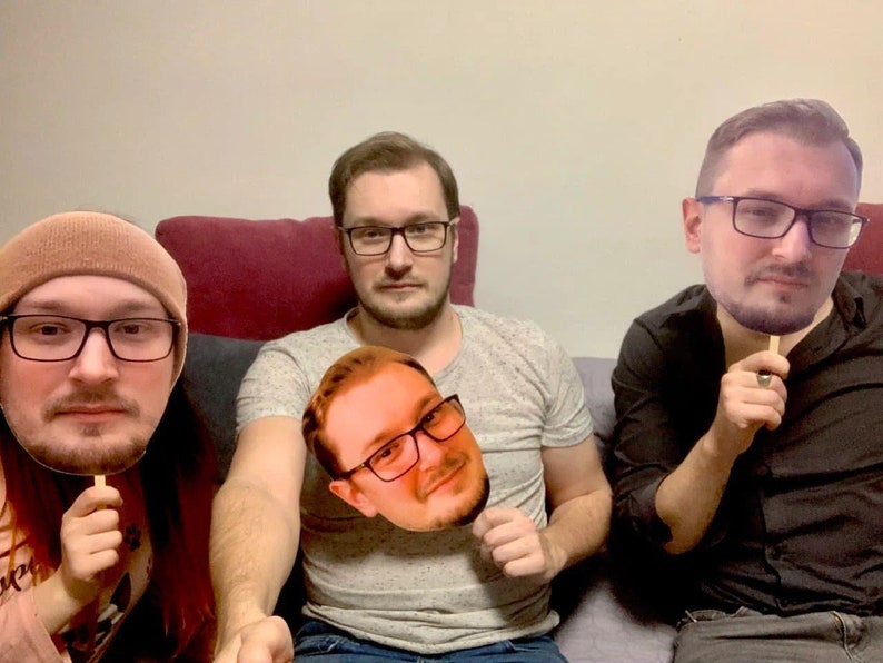 May include: Three people are holding up cardboard cutouts of their faces. The person on the left is wearing a brown beanie and a black shirt. The person in the middle is wearing a gray shirt and jeans. The person on the right is wearing a black shirt and jeans.