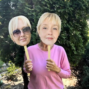 May include: Two cardboard face cutouts of a woman with blonde hair. The cutouts are held by a person wearing a pink shirt. The cutouts are on sticks.