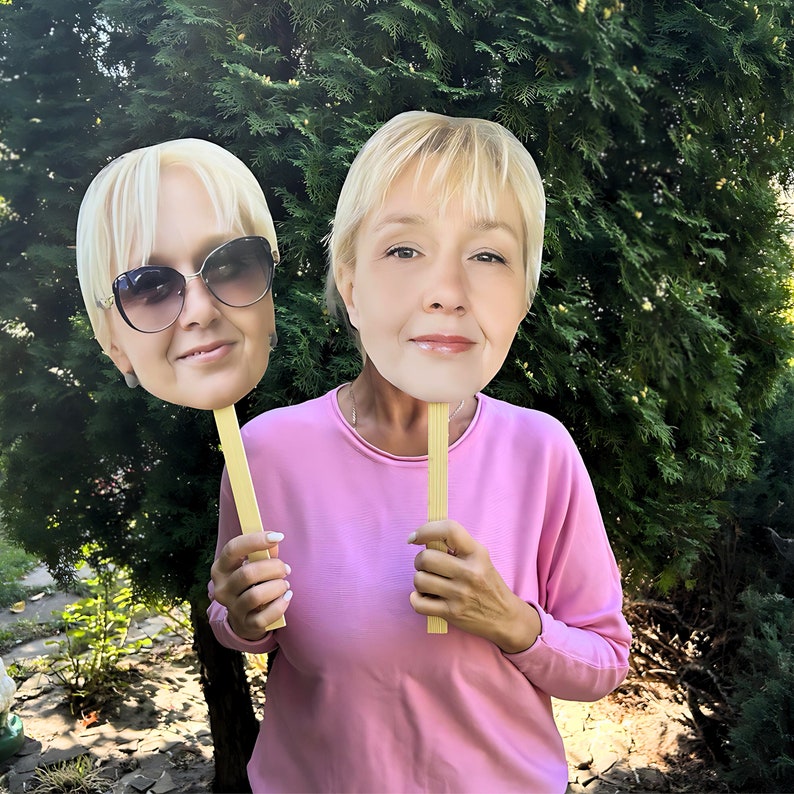 May include: Two cardboard face cutouts of a woman with blonde hair. The cutouts are held by a person wearing a pink shirt. The cutouts are on sticks.
