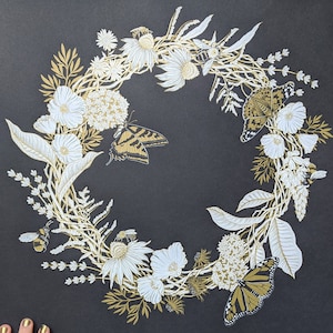 Peut inclure: Une couronne florale noire et dorée avec des papillons et des abeilles. La couronne est faite de fleurs, de feuilles et de vignes. Les papillons sont en vol et les abeilles bourdonnent autour des fleurs.
