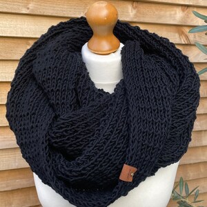 May include: A black knitted infinity scarf on a mannequin. The chunky scarf is made from textured yarn and features a small tag with a button. The scarf is designed to be worn around the neck and shoulders.