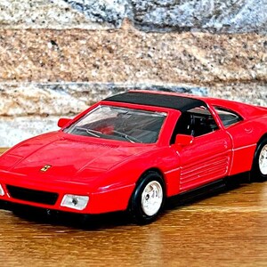 Puede incluir: Un coche de juguete rojo con techo negro y ruedas blancas. El coche tiene un diseño elegante y aerodinámico y un pequeño logotipo de Ferrari en el capó. El coche mide aproximadamente 15 cm de largo.