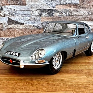 Op de afbeelding: Een zilvergrijze Jaguar E-Type roadster met een zwart kenteken dat "265 WK" aangeeft. De auto staat geparkeerd op een houten vloer voor een stenen muur.