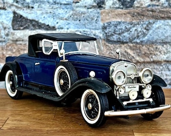 May include: A dark blue vintage convertible car model with black fenders and a black convertible top. The model has white-walled tires and chrome detailing, including headlights and a front bumper. The car is a classic collectible.
