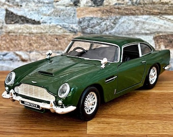 以下が含まれることがあります： シルバーのアクセントが施された濃い緑色のモデルカー。フロントグリル、バンパー、サイドミラーなど。クラシックなデザインで、ナンバープレートには「DBM 521V」と記載。木製の表面に置かれています。