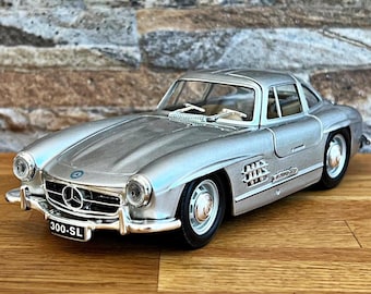May include: A silver 300 SL Mercedes-Benz coupe model car. The car has a black interior and a chrome grill. The car is sitting on a wooden surface.