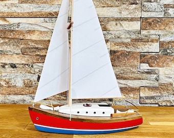 May include: A red and blue wooden model sailboat with white sails. The sailboat has a wooden mast and a white sail with a striped pattern. The sailboat is sitting on a wooden surface.