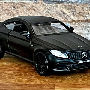 Op de afbeelding: Zwart speelgoedautomodel van een Mercedes-Benz AMG coupé. De auto heeft een zwarte grille met een zilveren Mercedes-Benz-logo en het woord "AMG" in zilveren letters.