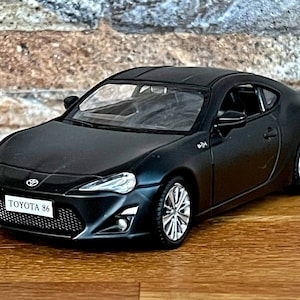 May include: A black Toyota 86 model car with a detailed front grille and headlights. The car is on a wooden surface.