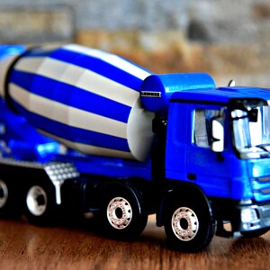 May include: Blue toy concrete mixer truck with a white and blue striped drum. The truck has a black grill and a black bumper.