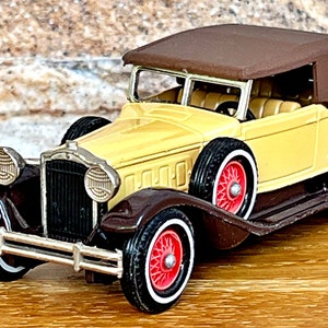 Könnte beinhalten: Ein vintage gelbes und braunes Spielzeugauto mit einem schwarzen Kühlergrill und roten Rädern. Das Auto hat ein braunes Stoffverdeck und ist ein Cabrio.