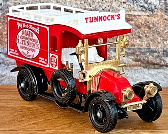 May include: A red and white toy delivery truck with a gold grill and black wheels. The truck has a white roof with the words "Tunnack's" printed on it. The side of the truck has a sign that reads "Just go to Tunnock's Baker & Confectioner J. Tunnock  Glasgow & Edinburgh Phone 28  Govanhill".